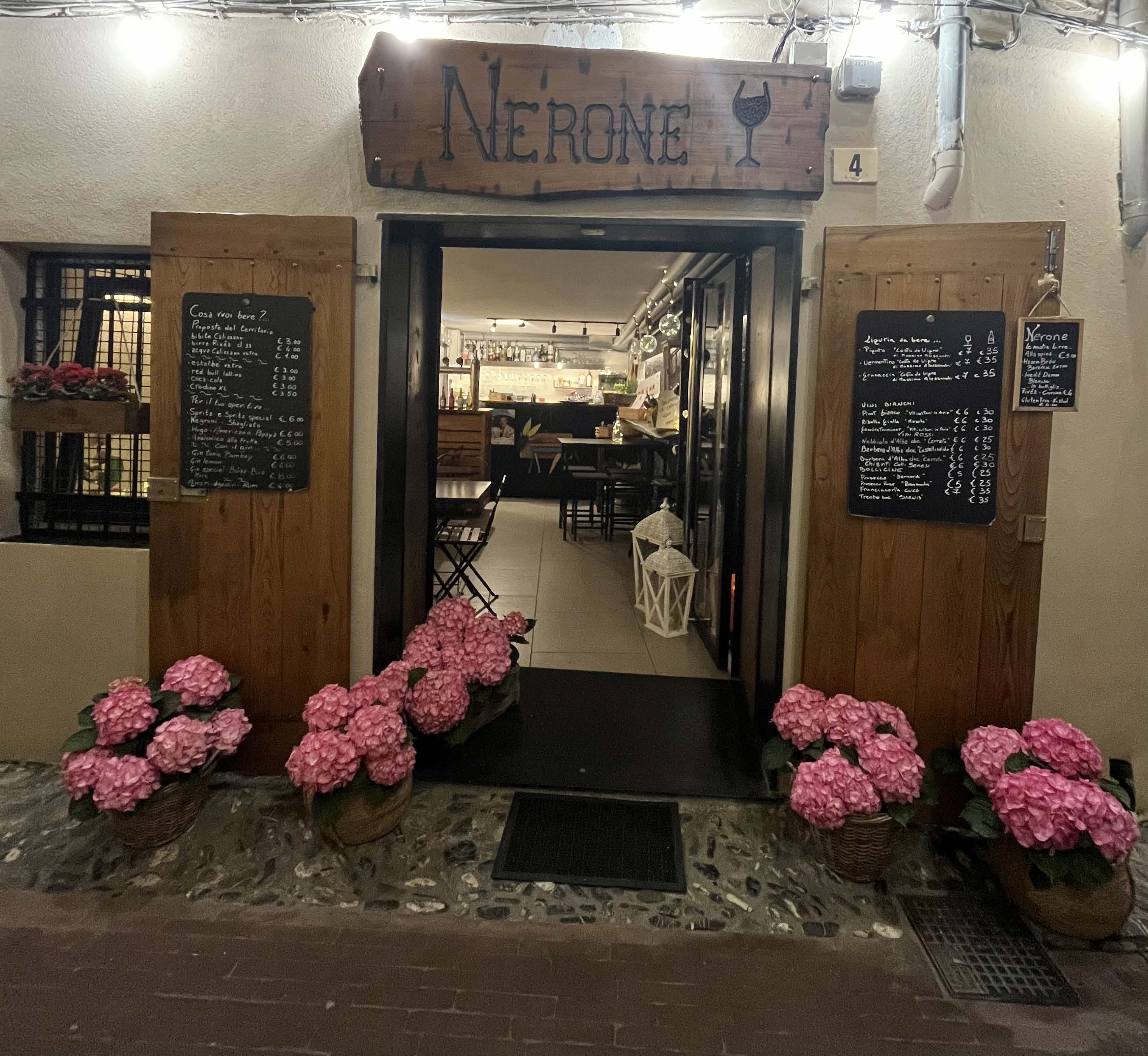 L'ingresso di Nerone nel centro storico di Albenga di sera: insegna in legno, porte aperte, fiori rosa